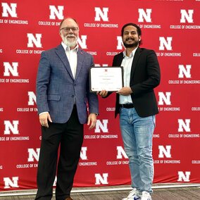 Two individuals pose on a red "College of Engineering" backdrop; one holds a certificate.