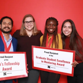 Group of four individuals holding award certificates against a solid red background.