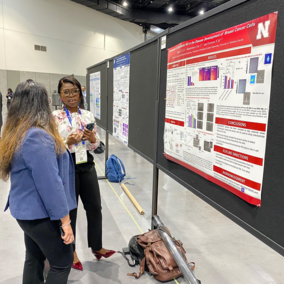 Two people discussing a research poster in a conference hall, with other attendees and posters nearby.