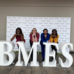 BMES letters displayed, four people posing behind, leaning on the letters in colorful outfits.